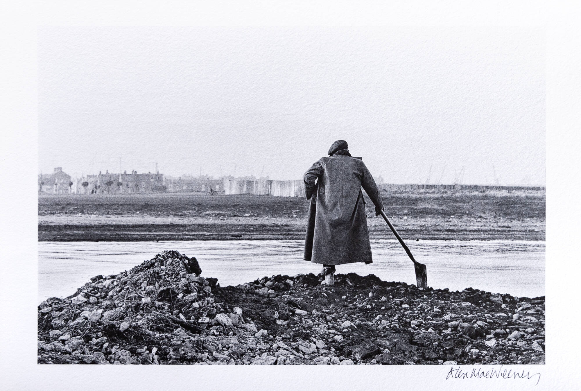 Black and white photograph of a man with a shovel standing in a rocky area with water in front of him. His back is to the viewer.